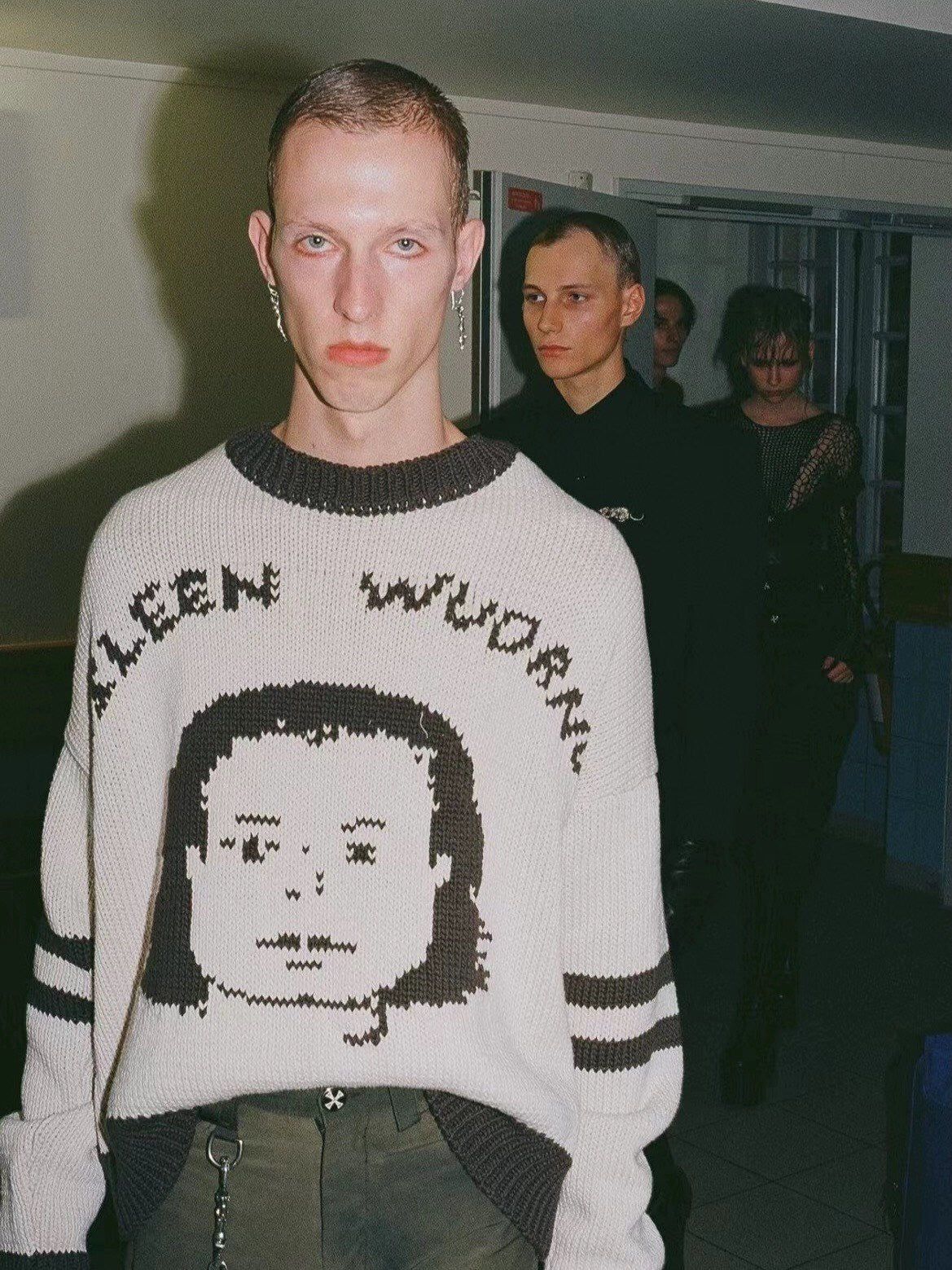 A model wearing a cream-colored knit sweater with a dark brown pixelated face graphic and the words 'ALEEN WOODS' across the chest, standing in a dimly lit indoor setting.