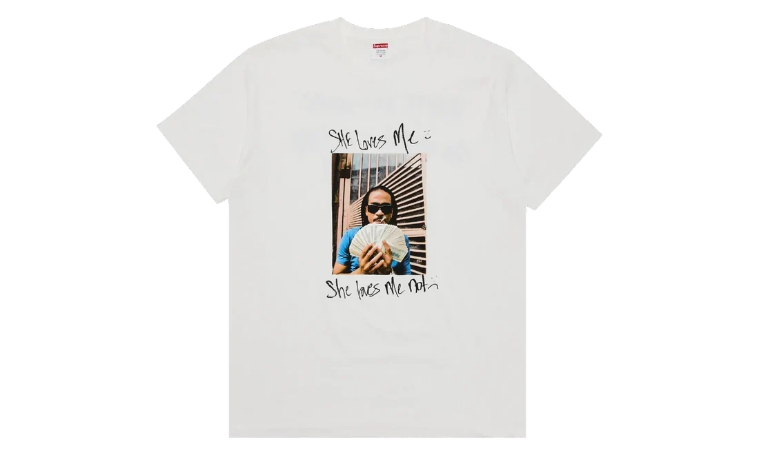 A white short-sleeve crewneck t-shirt featuring a graphic print on the front. The print includes a central rectangular photo of a person holding a fan, with the handwritten text 'She loves me' above and 'She hates me' below.